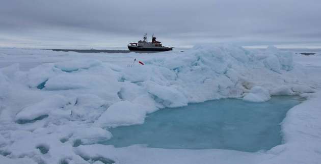 Foto eines Schmelztümpels auf einer Eisscholle. Im Bildhintergrund erkennt man den deutschen Forschungseisbrecher Polarstern. 